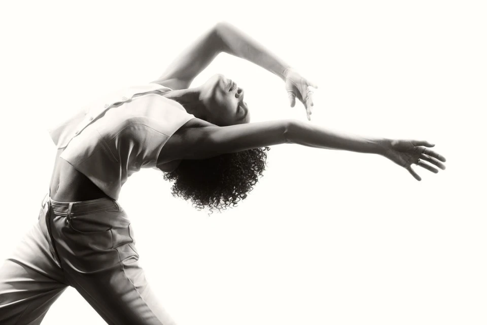 Artistic black and white studio portrait of a woman in motion wearing a crop top and pants against a bright white background.